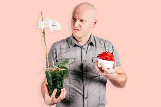 Handsome frowning bald middle-aged man in gray shirt hold white flowers in flowerpot and white gift box with red ribbon bow isolated on pink background. Valentine's day, dislike, emotion concept