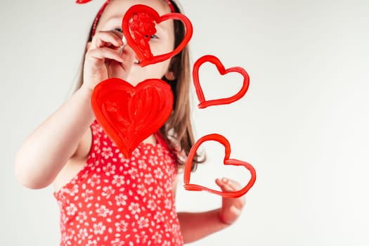 Little girl with long curly hair in red clothes draw hearts with paint on transparent glass smiling happily on white background. Family, love, holidays, birthday, Mother's day concept. Copy space.