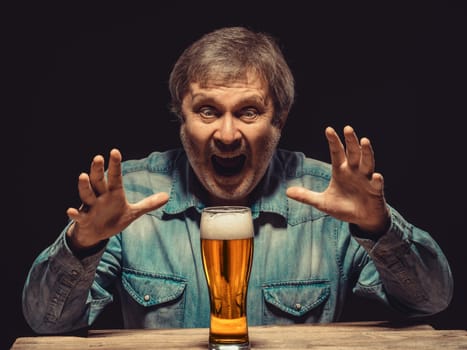 Enjoying his favorite beer.  The front view of handsome screaming man as fan in denim shirt with glass of beer, sitting at the wooden table. Concept of enthusiasm and ecstasy