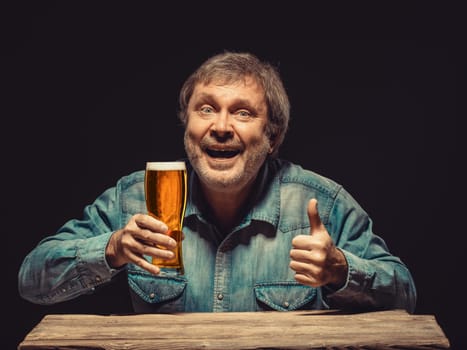 Enjoying his favorite beer.  The front view of handsome smiling  man as fan in denim shirt with glass of beer, sitting at the wooden table