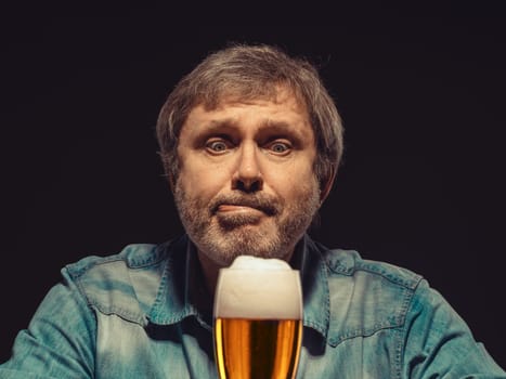 Enjoying his favorite beer.  The front view of handsome spellbound man in denim shirt with glass of beer, sitting at the wooden table. Concept of desire and love for beer