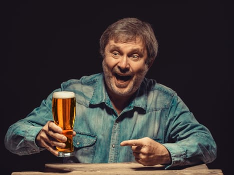 Enjoying his favorite beer.  The front view of handsome smiling  man as fan in denim shirt with glass of beer, sitting at the wooden table