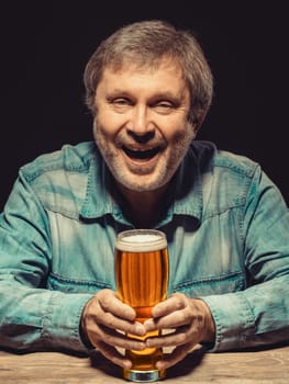 Enjoying his favorite beer.  The front view of handsome smiling  man as fan in denim shirt with glass of beer, sitting at the wooden table