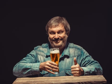 Enjoying his favorite beer.  The front view of handsome smiling  man as fan in denim shirt with glass of beer, sitting at the wooden table