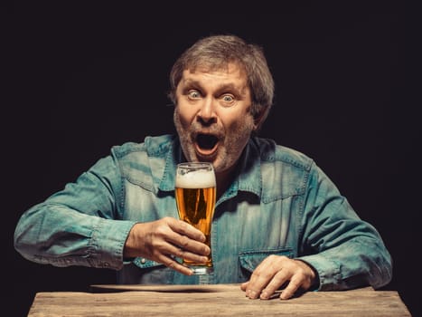 Enjoying his favorite beer.  The front view of handsome screaming man as fan in denim shirt with glass of beer, sitting at the wooden table. Concept of enthusiasm and ecstasy