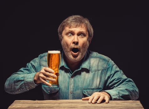 The front view of handsome man as enchanted and emotional fan in denim shirt with glass of beer, sitting at the wooden table. Concept of  surprise and exciting spectacle