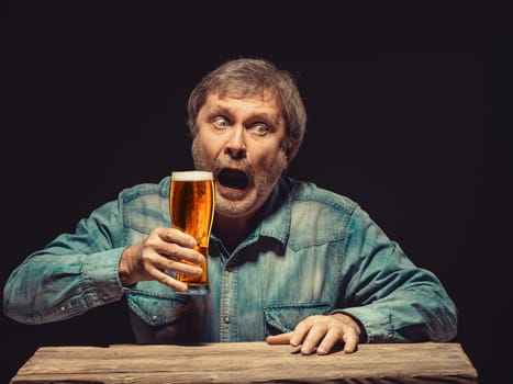 The front view of handsome man as enchanted and emotional fan in denim shirt with glass of beer, sitting at the wooden table. Concept of  surprise and exciting spectacle