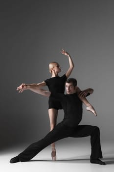 The two young modern ballet dancers in black suits posing over gray studio background