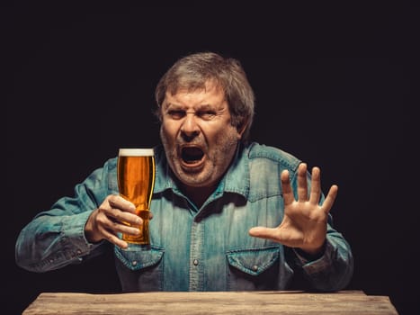 The front view of handsome man as emotional fan in denim shirt with glass of beer, sitting at the wooden table. Concept of fierce fan