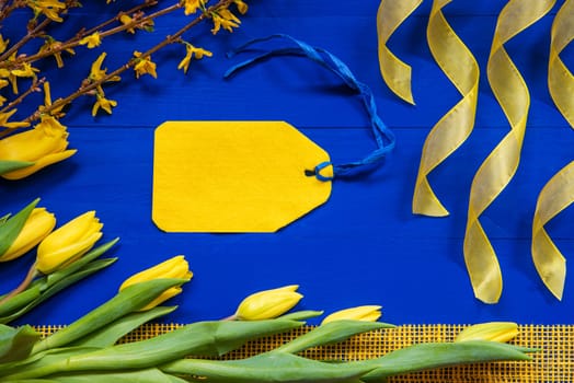 Yellow Label With Copy Space. Spring And Easter Decoration. Yellow Tulip Flowers, Branch And Ribbon. Blue Wooden Background