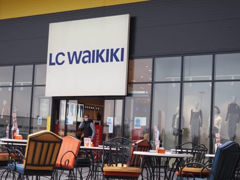 Sremska Mitrovica, Serbia, February 27, 2021 Shopping center. Clothing store LC WAIKIKI. A man wearing a surgical protective mask walks out of a shop's sliding doors. Cafe with metal chairs and tables