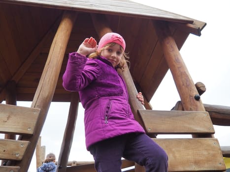 girl 7 years old on the playground. The child is dressed in warm demi-season clothes - a purple jacket and a pink hat. Wooden playground for climbing. Girl waving right hand.