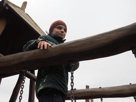 boy 8 years old on the playground. The child is dressed in warm, demi-season clothes - a brown hat and a green jacket. Children's wooden climbing area. Boy looking away.