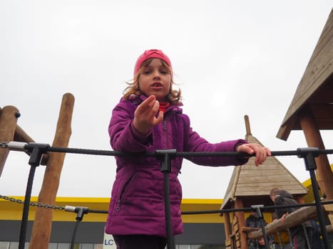 girl 7 years old on the playground. The child is dressed in warm demi-season clothes - a purple jacket and a pink hat. Climbing playground for children. The girl stretches out her right hand forward.
