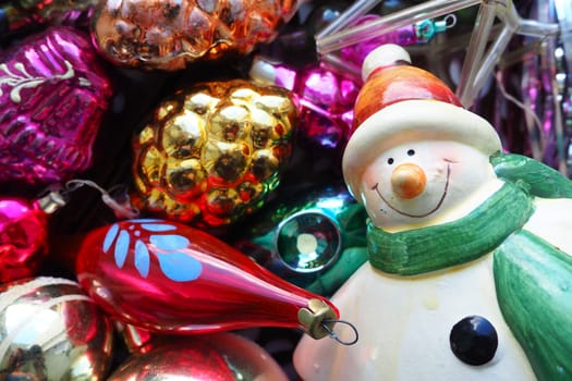 Cheerful smiling ceramic snowman in a green scarf and winter hat. New Year and Christmas decorations, tinsel. Multi-colored balls in the form of cones. Festive interior decor. New Year or Christmas.