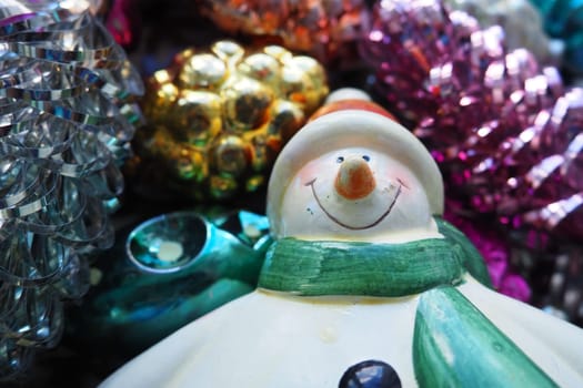 Cheerful smiling ceramic snowman in a green scarf and winter hat. New Year and Christmas decorations, tinsel. Multi-colored balls in the form of cones. Festive interior decor. New Year or Christmas.