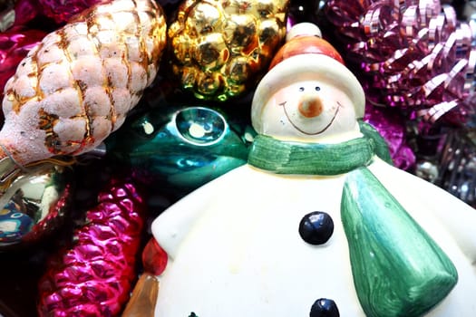 Cheerful smiling ceramic snowman in a green scarf and winter hat. New Year and Christmas decorations, tinsel. Multi-colored balls in the form of cones. Festive interior decor. New Year or Christmas.