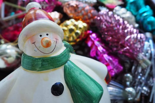 Cheerful smiling ceramic snowman in a green scarf and winter hat. New Year and Christmas decorations, tinsel. Multi-colored balls in the form of cones. Festive interior decor. New Year or Christmas.