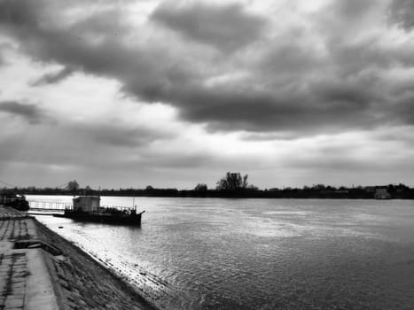 River landscape with a lonely ship at anchor. Cloudy weather with heavy clouds. Reflection of clouds in the water. The opposite bank is the horizon. Sava river, bank of Sremska Mitrovica, Serbia