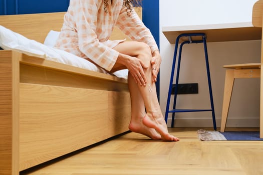 Unrecognizable woman applying body lotion on her legs at her bedroom