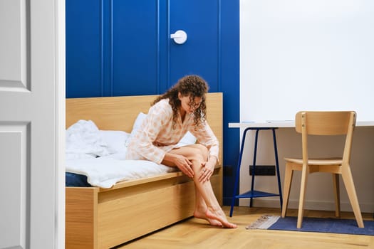 Young woman in bathrobe sitting in bedroom and rubbing cream on her legs