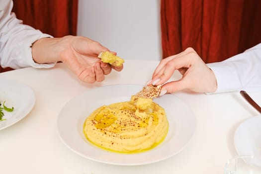 Two female hands dipping pieces of cracker into bowl with hummus