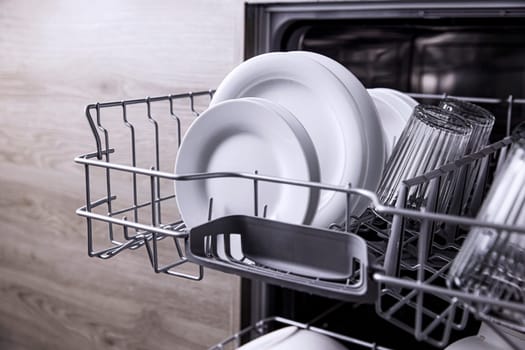 Front view of open automatic stainless steel built-in fully integrated top control dishwasher range machine with clean utensils, cutlery, glasses, dishes, plates inside on the upper deck kitchen