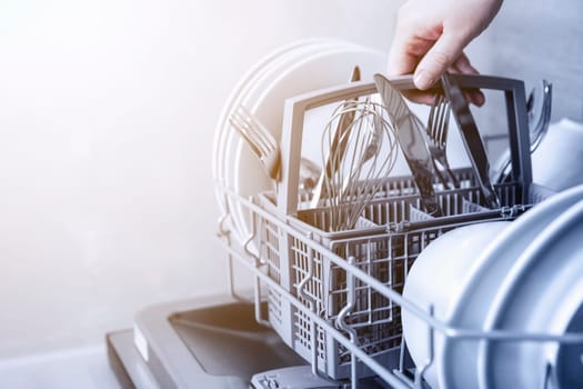 Close-up of female hand holding full cutlery basket with clean knife, fork, whisk. Loading to, empty out or unloading from open automatic dishwasher machine with clean utensils in home kitchen