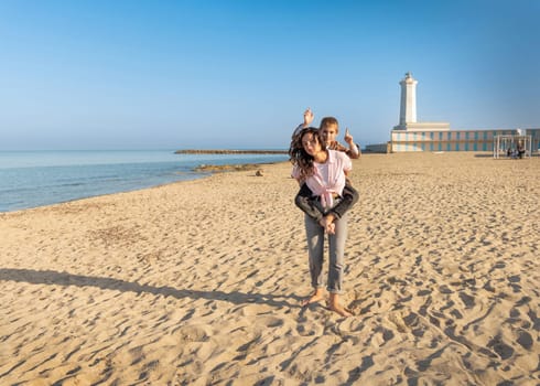 Happy Mother and son running on beach barefoot in a sunny day against the lighthouse. Toddler kid boy spending time together with his Mom and having fun near the sea at sunset