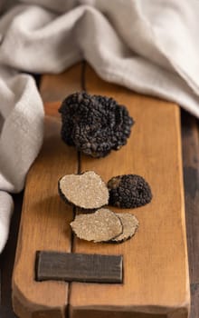 Whole and sliced black truffles mushroom on wooden board on dark brown table near grey napkin, close up. Culinary delight ingredient.
