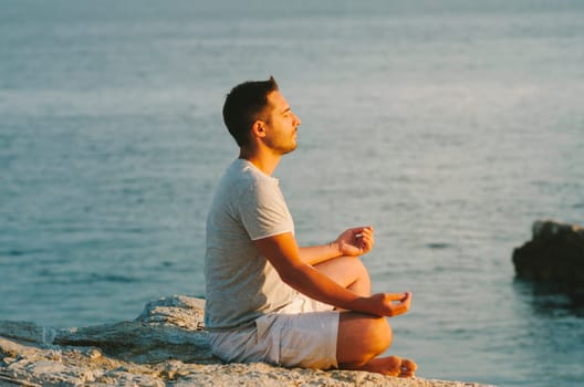 Young man sitting on the sea coast, relaxing and meditating by the water at sunset. Meditation and sun gazing in nature.
