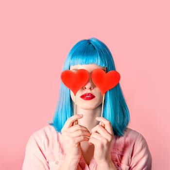 Young stylish woman with blue hairstyle and red lipstick holding valnetine's heart on her eyes. Hipster funny girl model over pink pastel studio background. Love and valentine's day design concept.
