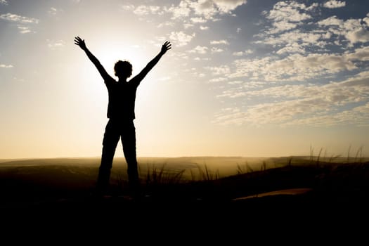Silhouette of person standing on a dune during sunset, arms raised. Copy space.