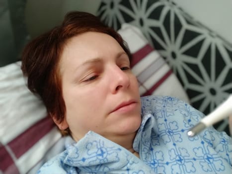 Sick woman in bed. Headache, runny nose and cold. Paleness of the face and faintness of the body. Feeling unwell. The patient lies on a pillow, holds a thermometer and measures the temperature