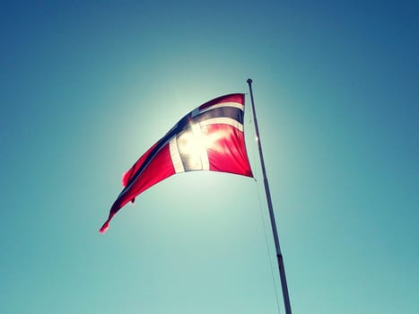 Flag of Norway waving in the wind, on a pole, against blue sky. Creative edit.