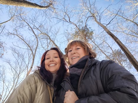 Two women stand together smiling happily in a winter forest, trees reaching towards the clear blue sky above them.