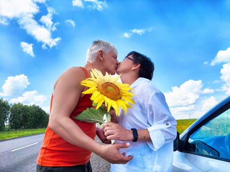 A middle-aged couple shares a sweet kiss by the roadside, surrounded by clear blue skies and fluffy clouds. The man holds a bright sunflower, symbolizing love and joy during their trip.