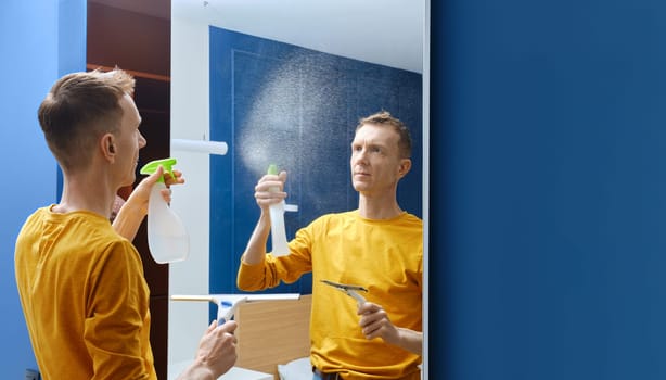 Middle-aged man spraying cleaning liquid on mirror at home to clean it