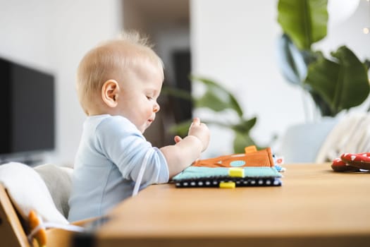 Happy infant sitting at dining table and playing with his toy in traditional scandinavian designer wooden high chair in modern bright atic home. Cute baby playing with toys.