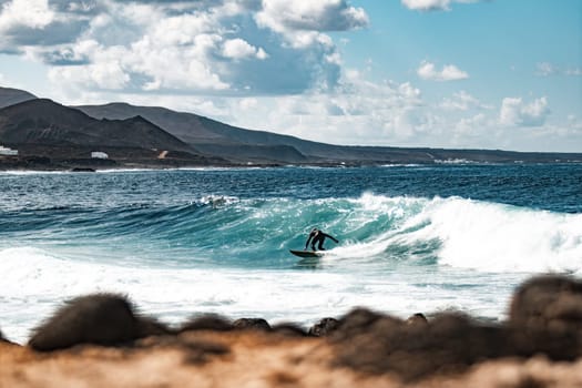 Wild rocky coastline of surf spot La Santa Lanzarote, Canary Islands, Spain. Surfer riding a big wave in rocky bay, volcano mountain in background