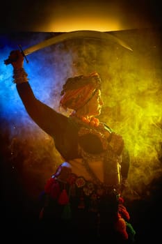 A woman in a colorful headscarf and jewelry poses With a saber in front of wall of colorful tassels and smoke around
