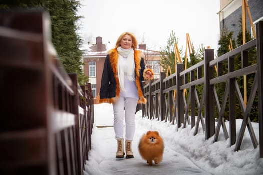 A middle-aged woman in winter attire enjoys a stroll with her fluffy, red Pomeranian dog along a snowy walkway.