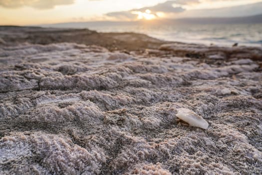 Sand and stones covered with crystalline salt on shore of Dead Sea, clear water background.