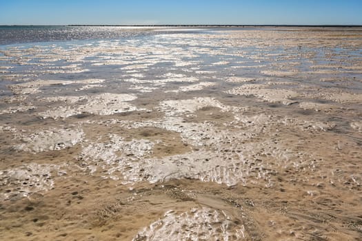 Sun shines to shallow sea during low tide, wet sand visible, sandy shore in distance.
