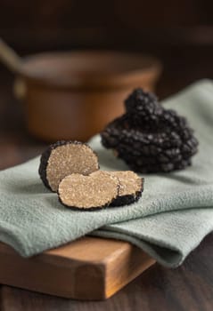 Whole and sliced black truffles mushroom on wooden board on green napkin on dark brown table, close up. Culinary delight ingredient.