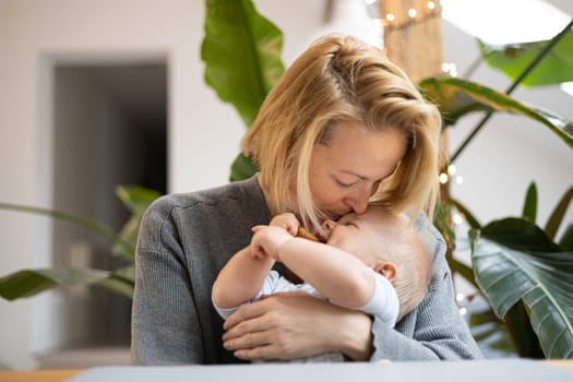 Portrait of young mother cuddling and kissing her adorable little baby boy while sitting at the table at home. Sensory stimulation for baby development