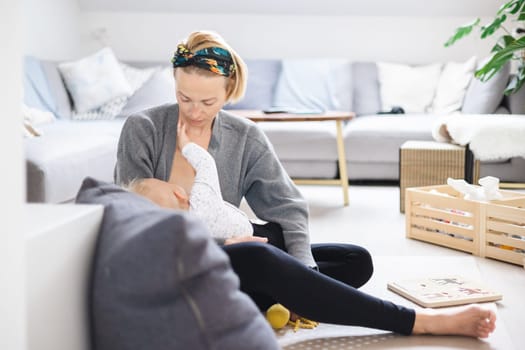 Young woman breastfeeding her infant baby boy casualy sitting on child's playing mat on living room floor at home