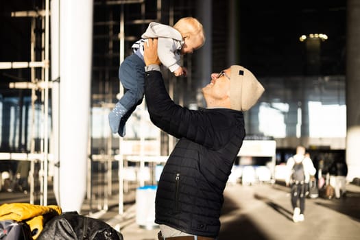 Father happily holding and lifting his infant baby boy child in the air after being rejunited in front of airport terminal station. Baby travel concept