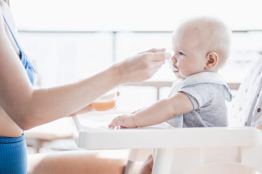 Mother spoon feeding her baby boy child in baby chair with fruit puree on a porch on summer vacations. Baby solid food introduction concept
