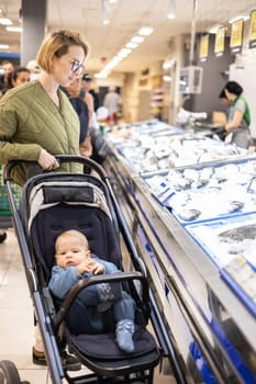 Casualy dressed mother choosing fish in the fish market department of supermarket grocery store with her infant baby boy child in stroller